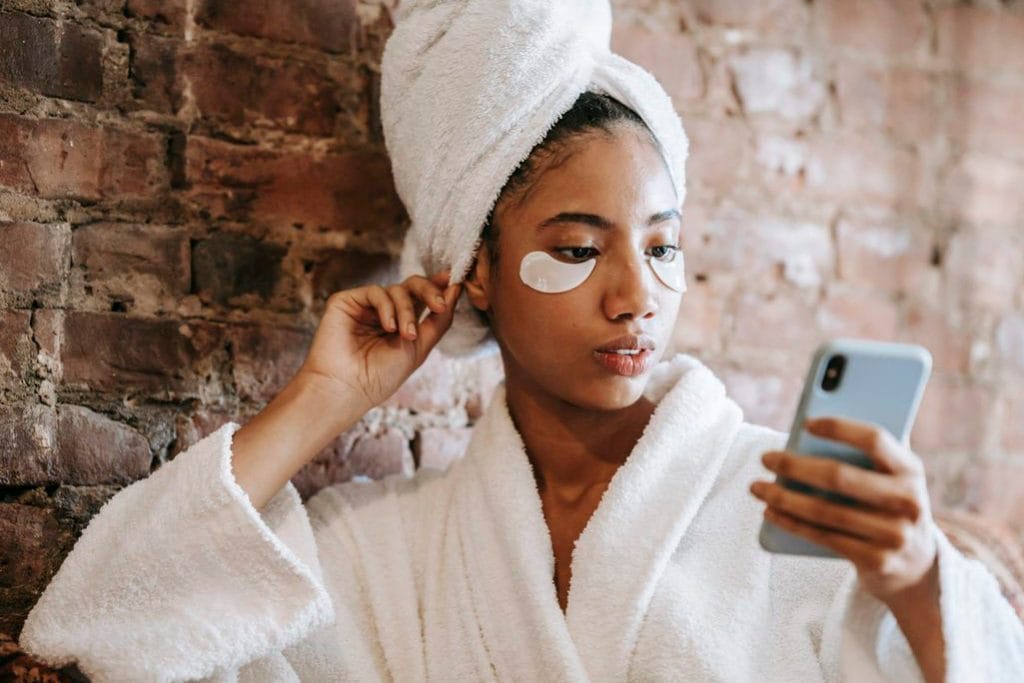 Ethnic woman in eye patches watching smartphone in spa salon